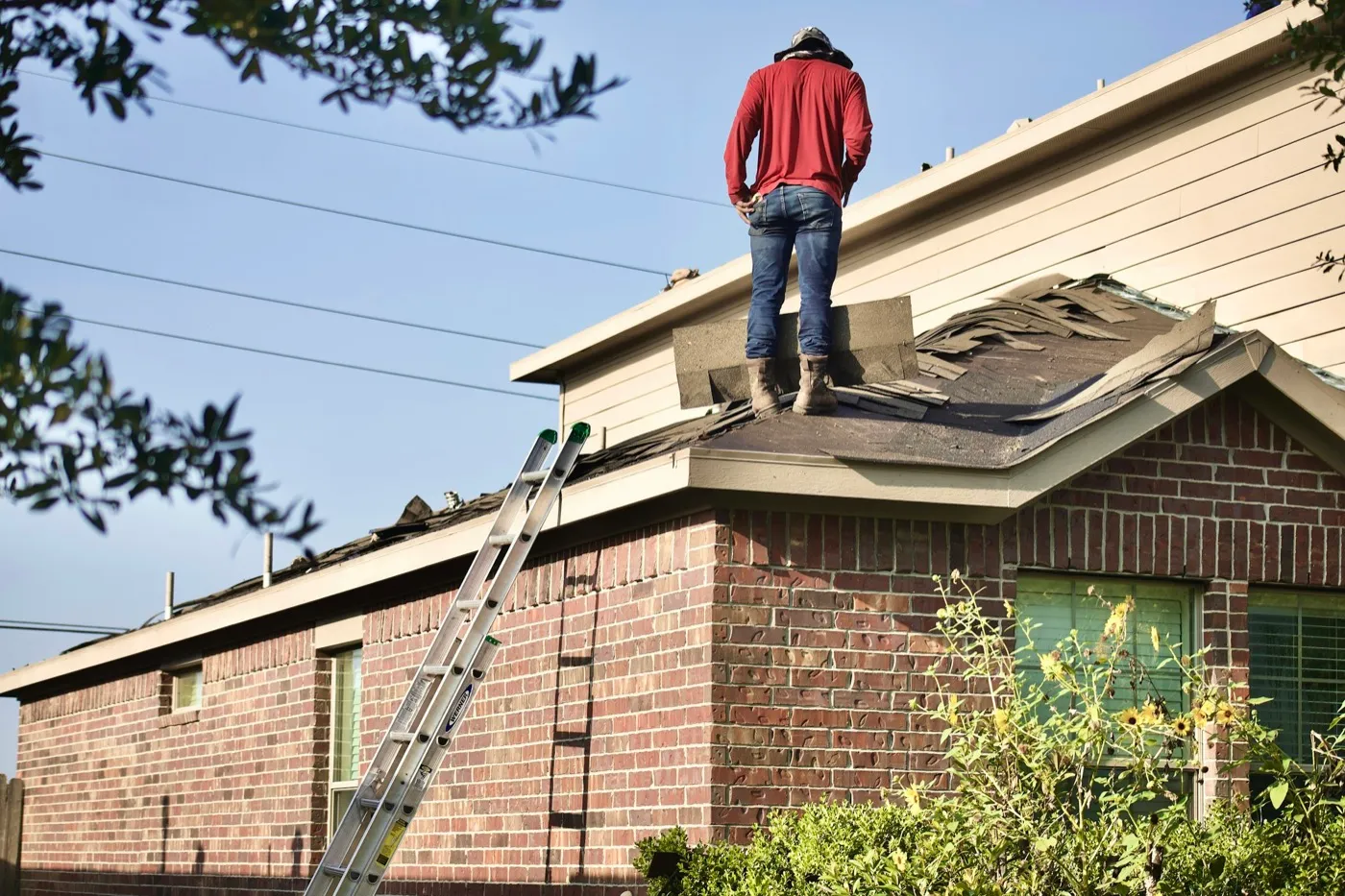 A roofer stands on the partly stripped roof of a one-story brick ranch house during a roof replacement, with torn-off shingles on the bare deck and an aluminum ladder against the eave.