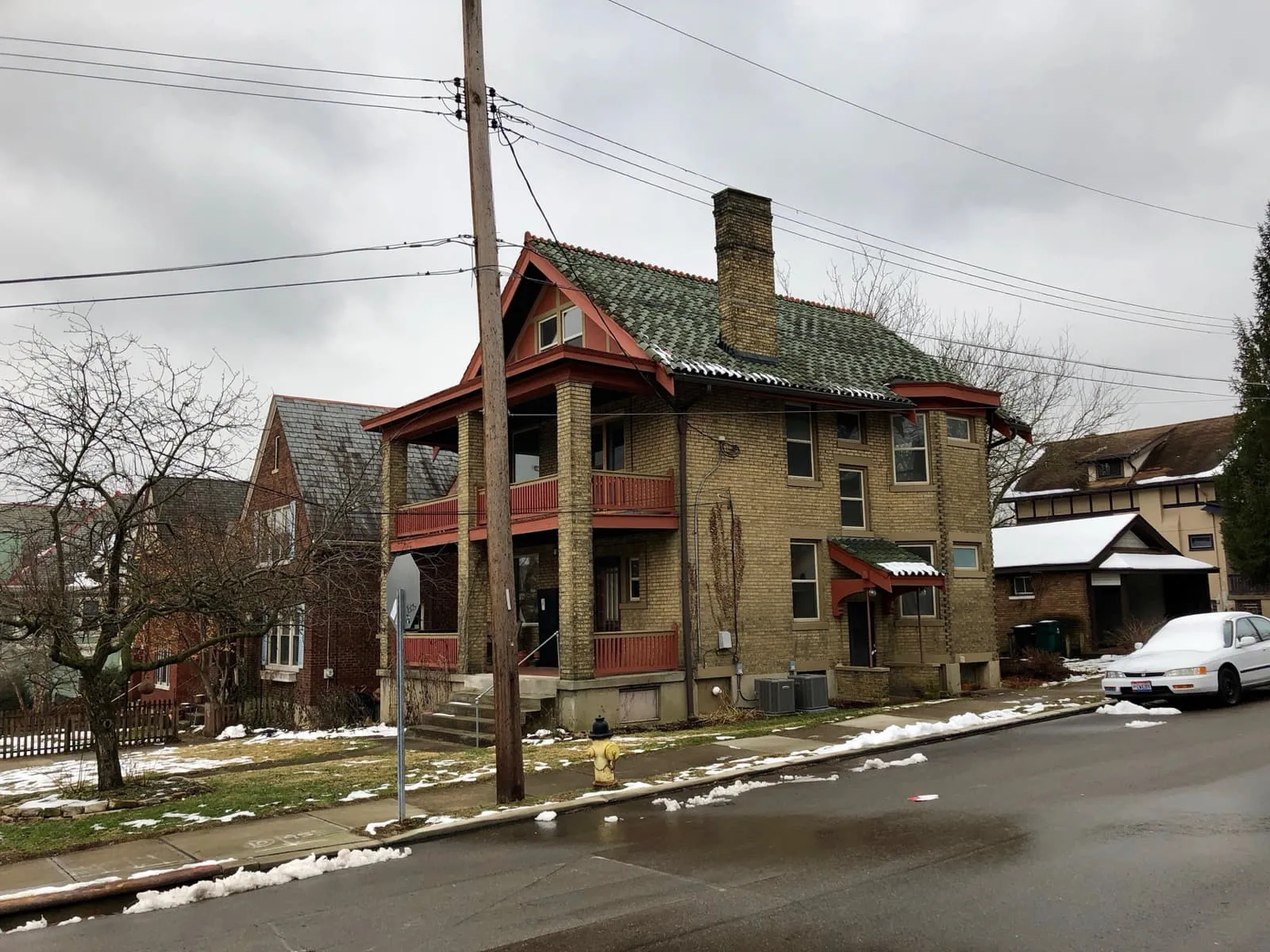 A yellow-brick Craftsman house with a green tile roof and a wide porch on Hamilton Avenue in Northside, Cincinnati, with snow on the ground and an air-conditioning condenser beside it.