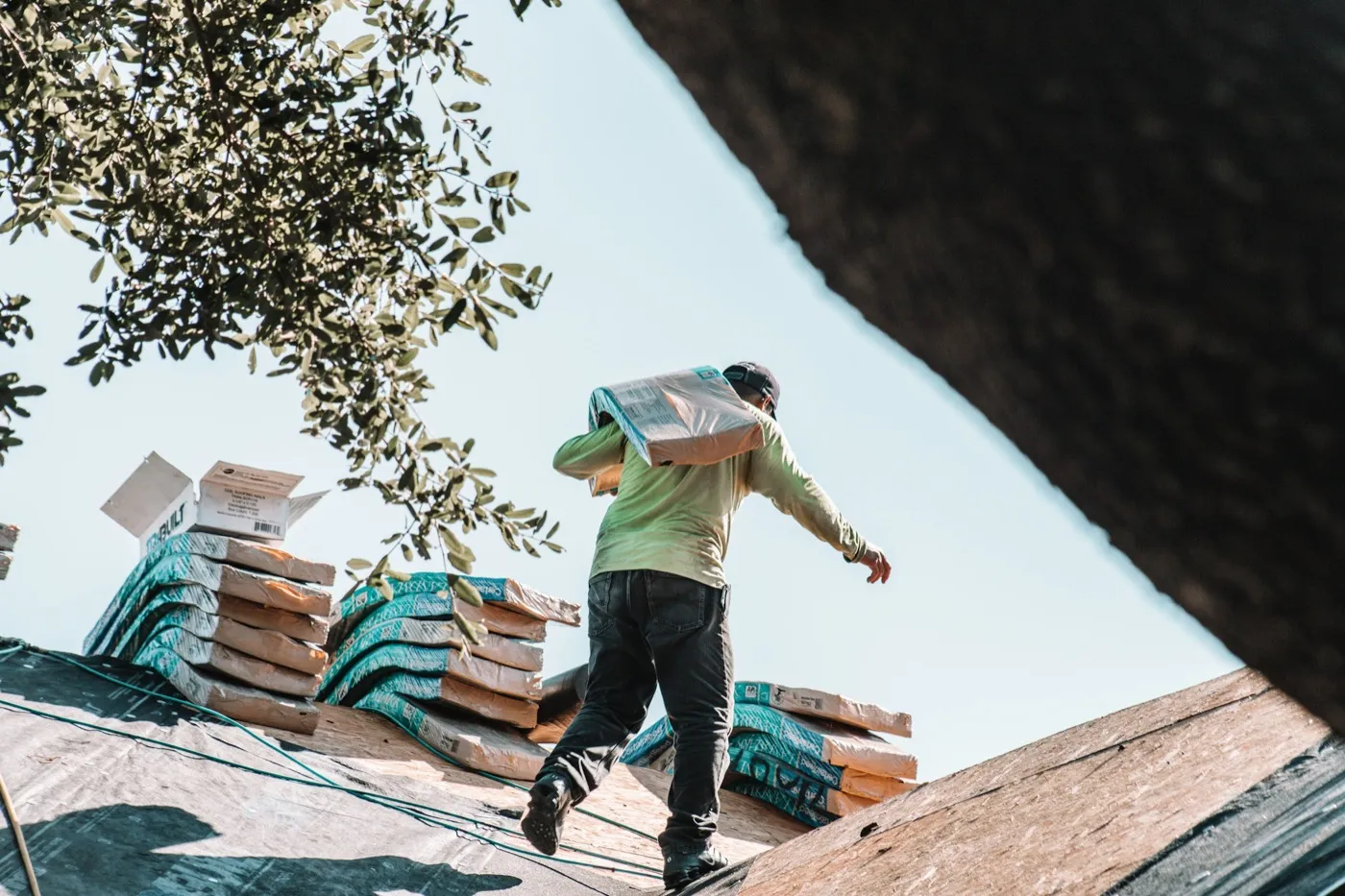 A roofer carries a bundle of asphalt shingles across freshly laid roof decking during a roof replacement, with more shingle bundles stacked along the roof.